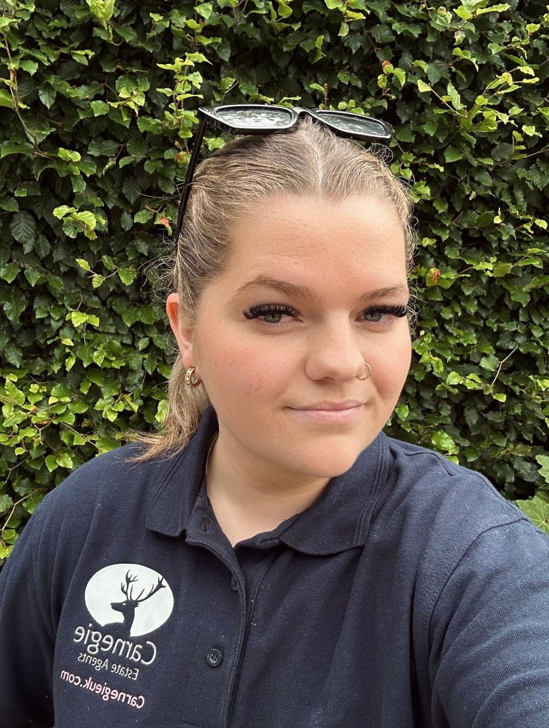 Person wearing a navy polo shirt with a logo, standing in front of a leafy green hedge.